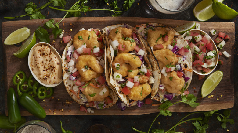 3 tacos filled with fried shrimp with dips, slices of lemon and garnish on top of a wooden board