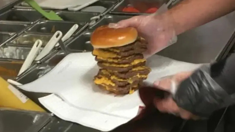 A fast food worker on a food line, holding and wrapping a burger with nine patties.