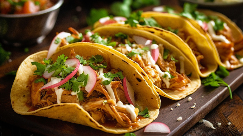 A row of chicken tacos on a board garnished with cilantro and radish slices.