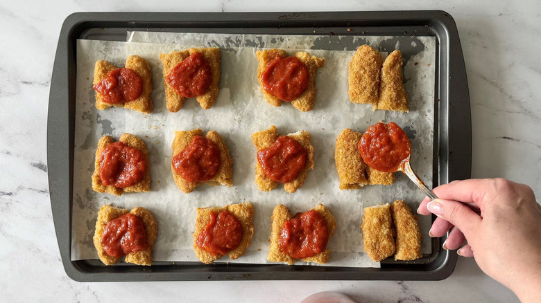 Spooning marinara sauce onto chicken strips on baking sheet