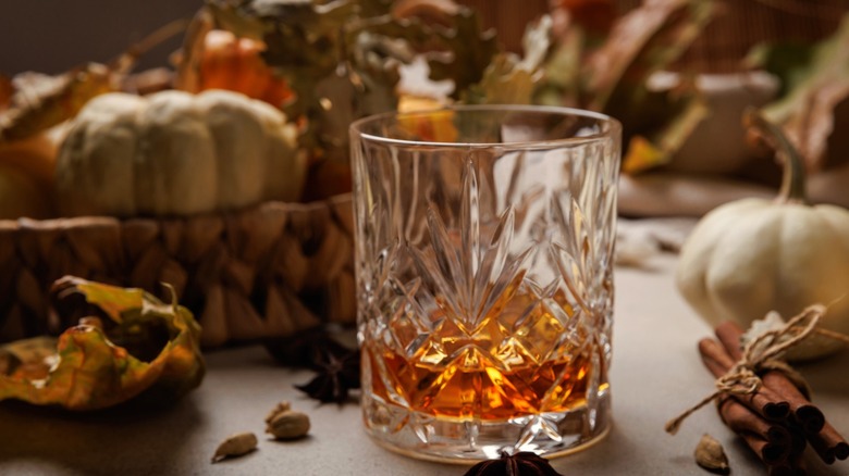 A rocks glass with a brown cocktail in it surrounded by small pumpkins and dried leaves