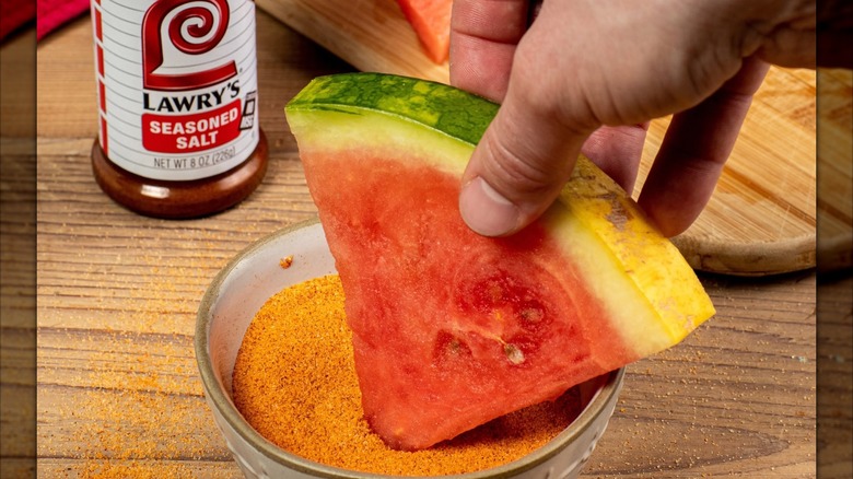 A hand dipping a watermelon slice into a bowl of Lawry's seasoned salt