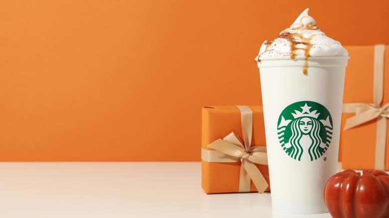 A venti Starbucks Pumpkin Spice Latte with whipped cream and topping, in an orange setting with orange gift boxes