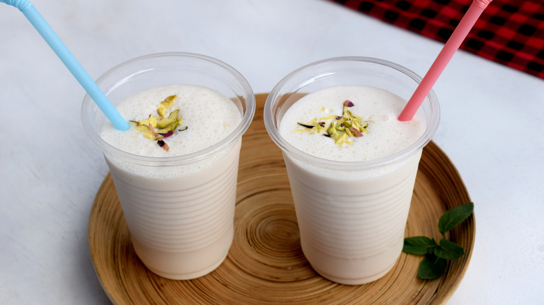 Two glasses of lassi topped with chopped pistachios on a wooden plate