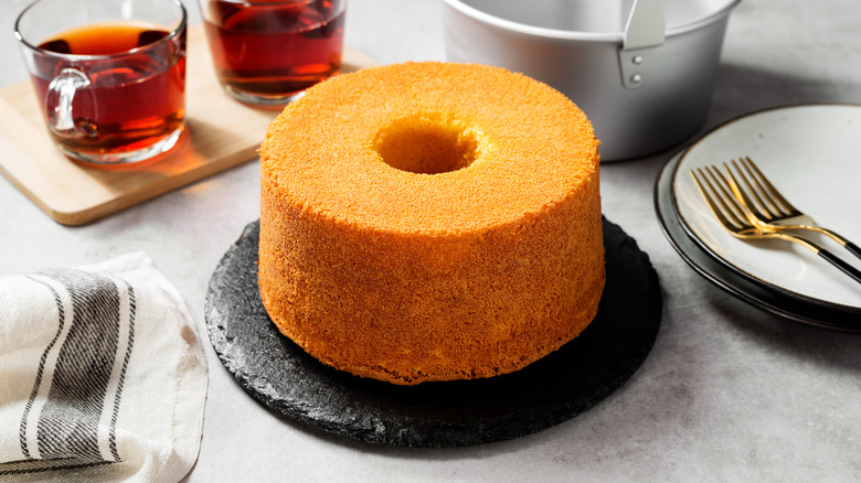 A round chiffon cake next to cups of tea on a table with plates, forks, and a towel.