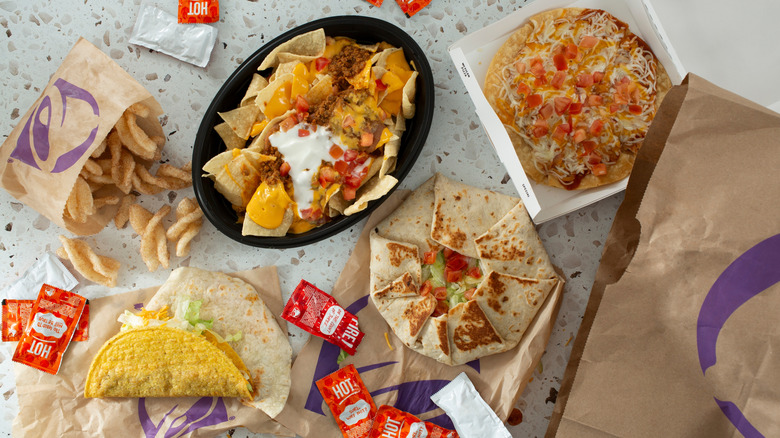 An array of Taco Bell items, including cinnamon twists, tacos, nachos, a Mexican Pizza, and a crunchwrap with various hot sauce packets on a counter
