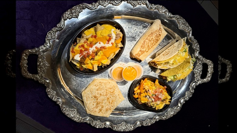 The Taco Bell Volcano Sauce menu on a silver platter, including crunchy taco, cheesy g, steak nachos, quesarito, crunchwrap, and BellGrande nachos
