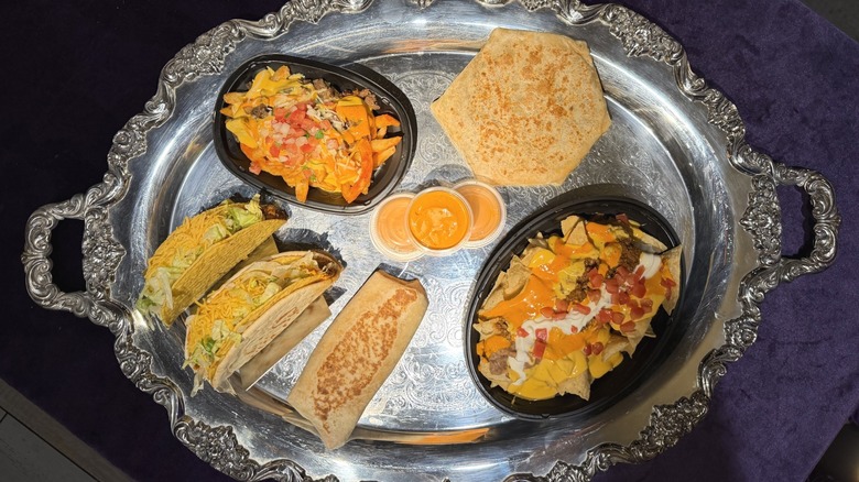 The Taco Bell Volcano Sauce menu on a silver platter, including crunchy taco, cheesy g, steak nachos, quesarito, crunchwrap, and BellGrande nachos