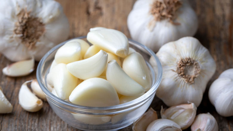 Garlic cloves in a glass bowl with whole garlic bulbs on the side, all on a wooden table