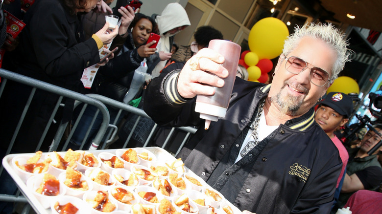 Guy Fieri holding a squeeze bottle of BBQ over a tray of samples of fried chicken