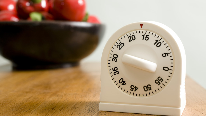 A white kitchen timer with black numbers and a black bowl of red apples in background
