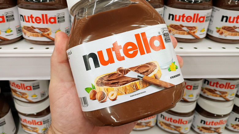 A hand holds a jar of Nutella while many jars sit on a grocery store shelf behind