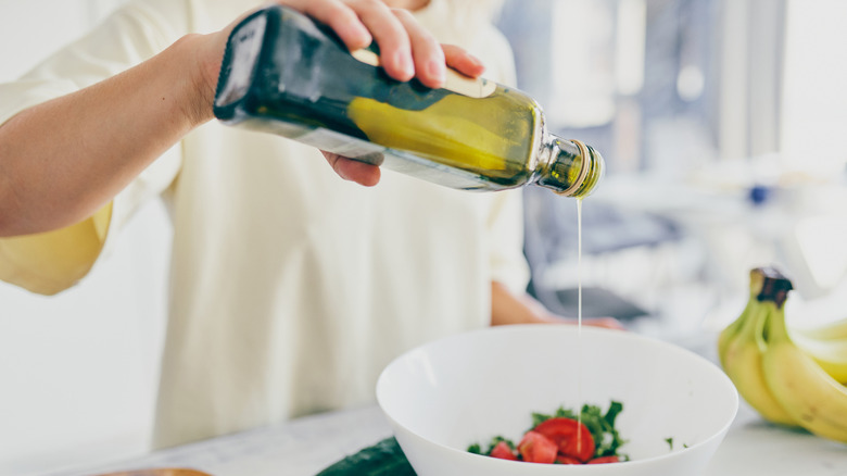 A person pours olive oil over tomatoes and greens in a white bowl