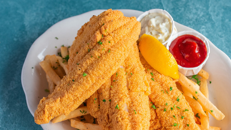 a plate with three pieces of fried fish, french fries, ramekins of tartar sauce and ketchup, and a lemon wedge