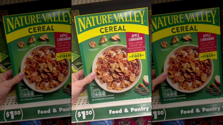 A hand holding a green box of Nature Valley Apple Cinnamon Breakfast Cereal