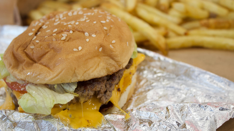 Closeup of a burger from Five Guys with melting cheese sticking to the foil wrapper and fries in the background