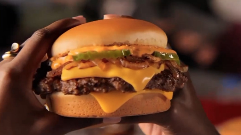Two hands holding a Smashed Jack cheeseburger, topped with sauce, pickles, and grilled onions