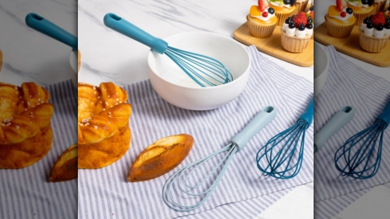 A blue whisk in a white bowl with two more blue whisks nearby on a striped tea towel with bread and pastries nearby.