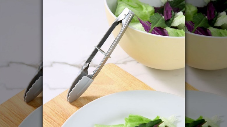Mini food tongs on a wooden cutting board by a salad bowl with with a salad plate nearby