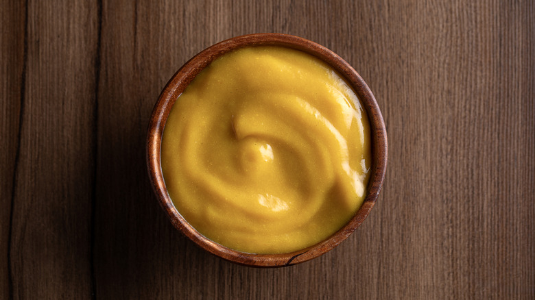 A small brown bowl of yellow mustard on a wooden background