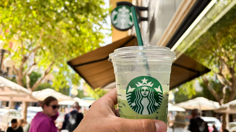 Hand holding a Starbucks drink in front of a Starbucks