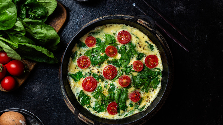 An omelet cooking in the pan with leaves of spinach and halved tomatoes