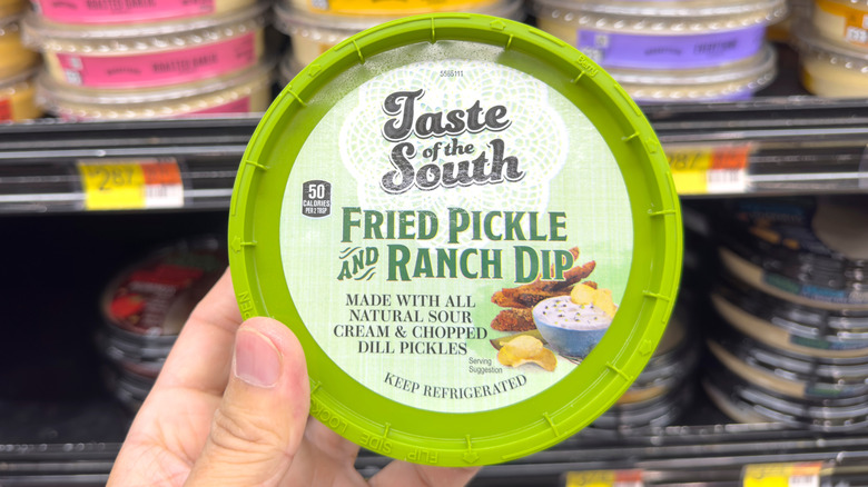 A hand holding Taste of the South fried pickle and ranch dip in front of a dip display in a grocery store