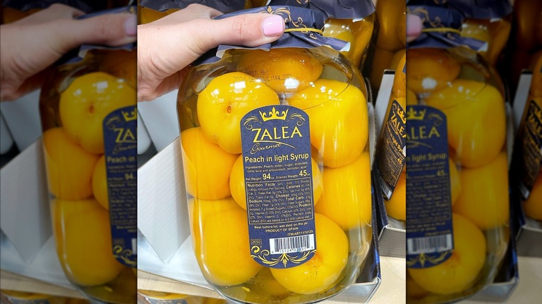 A hand holding a large clear glass jar of Zalea gourmet peaches in light syrup