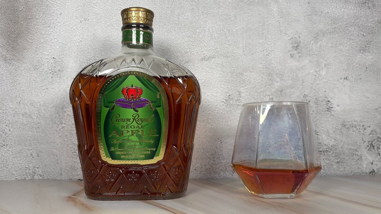 Bottle of Crown Royal apple whiskey next to glass of whiskey on grey background