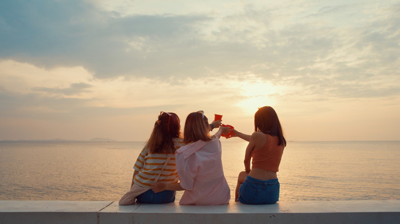 Three women toasting with red cups by water with sunset in distance