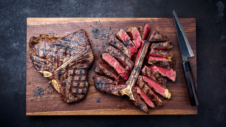 two grilled porterhouse steaks, one intact and one sliced for serving, resting on a wooden board next to a steak knife