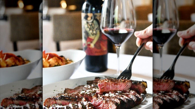 a hand sticks a fork into a porterhouse steak that is plated and sliced, with a glass of red wine accompanying