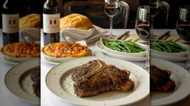 a porterhouse steak on a plate, surrounded by family-style side dishes of green beans, potatoes, and bread, with glasses of red wine