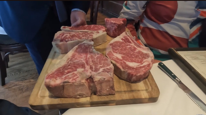 a server shows diners selections of raw steak on a wooden board, with the porterhouse cut front and center of frame