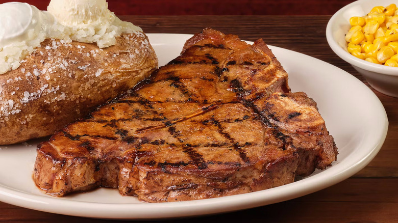a porterhouse steak with diamond hatch marks on a plate with buttery baked potatoes and a side bowl of corn