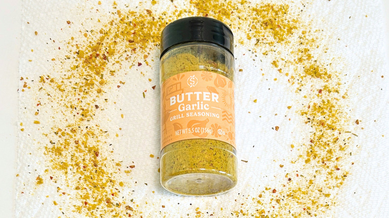 A bottle of Stonemill Butter Garlic Grill Seasoning surrounded by butter garlic seasoning on a white background