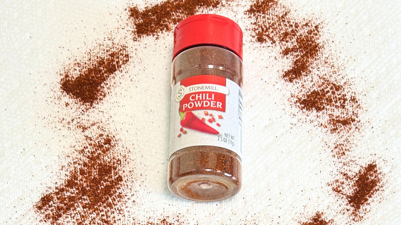 A container of Stonemill Chili Powder surrounded by chili powder on a white background
