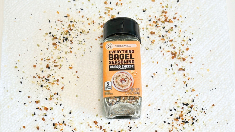 A bottle of Stonemill Everything Bagel Seasoning Asiago Cheese next to everything bagel seasoning on a white background