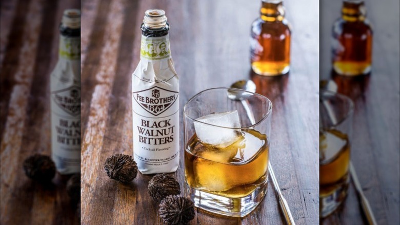A bottle of Fee Brothers' black walnut bitters on a table with black walnuts and a whiskey cocktail