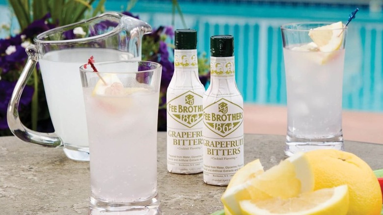 Two bottles of Fee Brothers' grapefruit bitters next to two glasses of clear cocktails and a bowl of sliced lemons on a table next to a pool