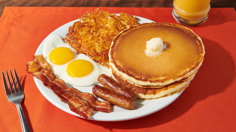 a plate of bacon, sausage links, sunny side up eggs, hash browns, and two pancakes on a table with an orange placemat