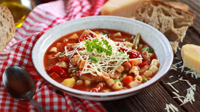 Minestrone soup in a white bowl next to a checkered cloth and chunks of cheese and bread