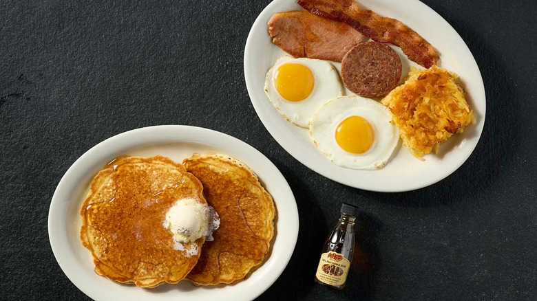 A plate of pancakes next to a plate with sunny-side up eggs, hashbrown casserole, sausage, and bacon, all on a black table with a bottle of syrup