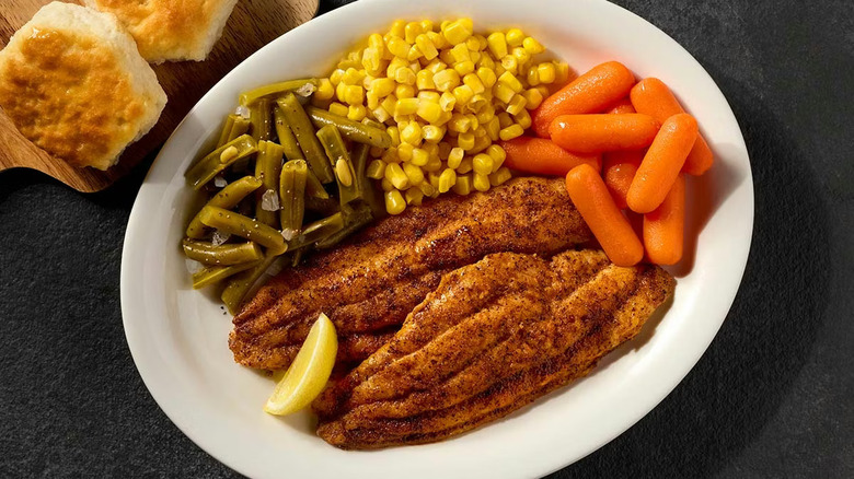 Grilled catfish on a white plate with carrots, corn, and green beans, all placed on a black table with biscuits on the side