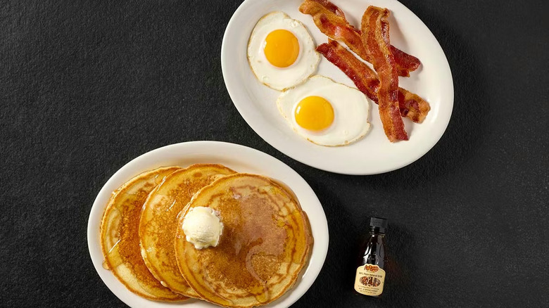 A plate of pancakes next to a plate with sunny-side up eggs and bacon, all on a black table with a bottle of syrup