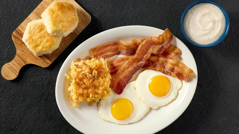 A plate full of bacon, sunny side up eggs, and hashbrown casserole on a black table with biscuits and gravy on the side