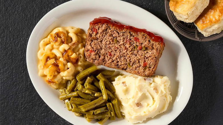 A plate of a slide of meatloaf, mashed potatoes, mac and cheese, and green beans on a black table with biscuits on the side