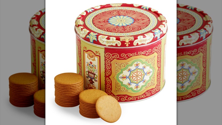 A red, gold, and white tin with floral designs, next to several thin ginger snaps