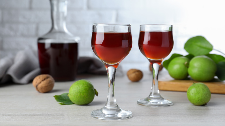 Two glasses of nocino, next to some fresh walnuts, with a bottle in the background