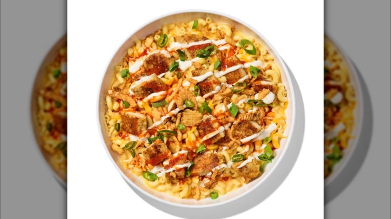 A bowl of mac and cheese with chicken, scallions, fried onions, ranch, and buffalo sauce drizzled over top against a white background